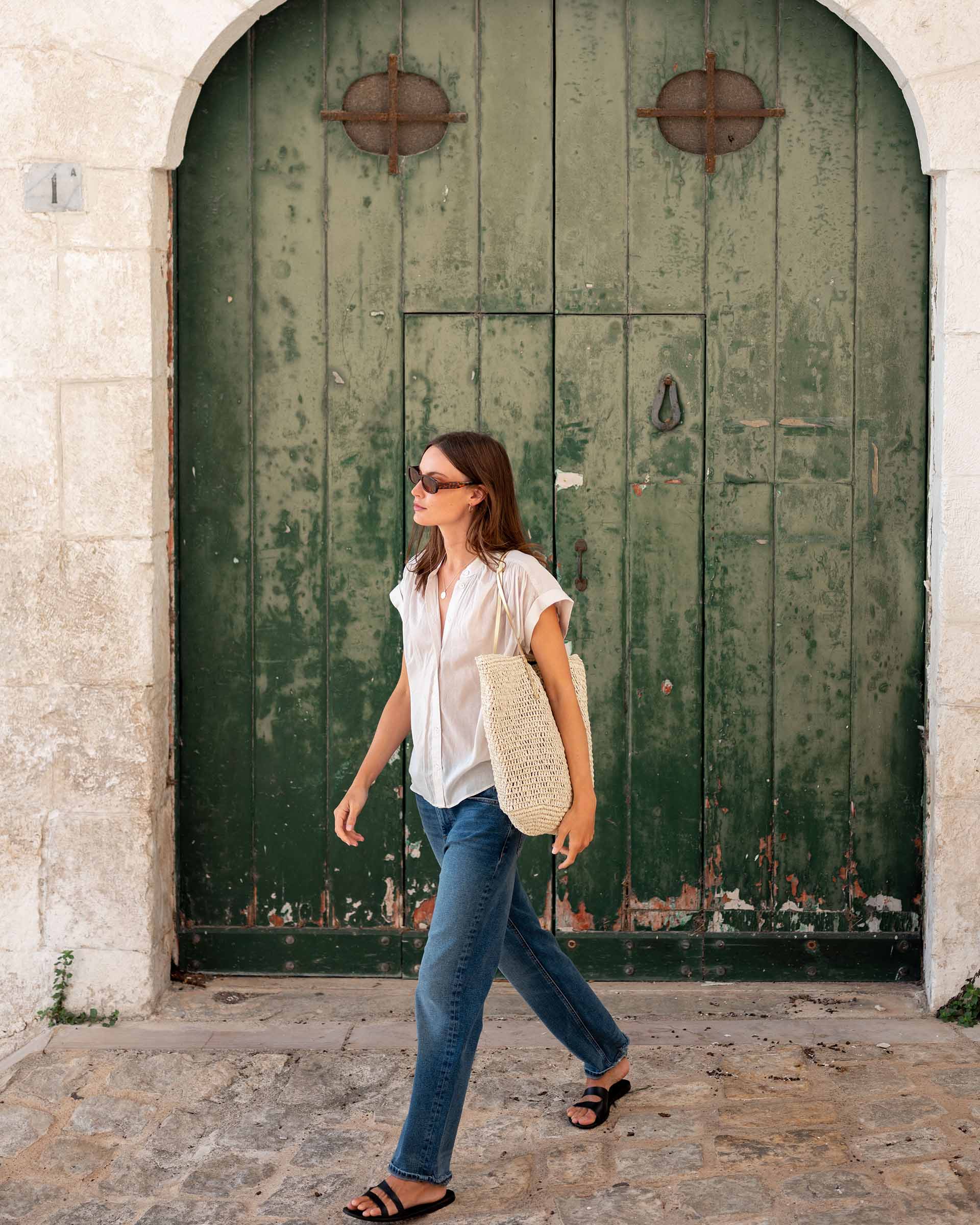woman walking away from green door wearing aphrodite button up shirt in moonbeam wearing sunglasses