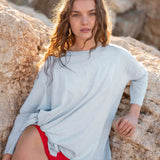 Female wearing a light blue oversize tee over a red bathing suit leans on rock.