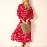 Woman wearing a red patterned dress holding a woven bag against a plain background