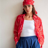 Woman wearing a red bandana, red shirt, white top, and blue pants against a plain background