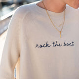 Person wearing a cream sweater with 'rock the boat' text and layered necklaces.