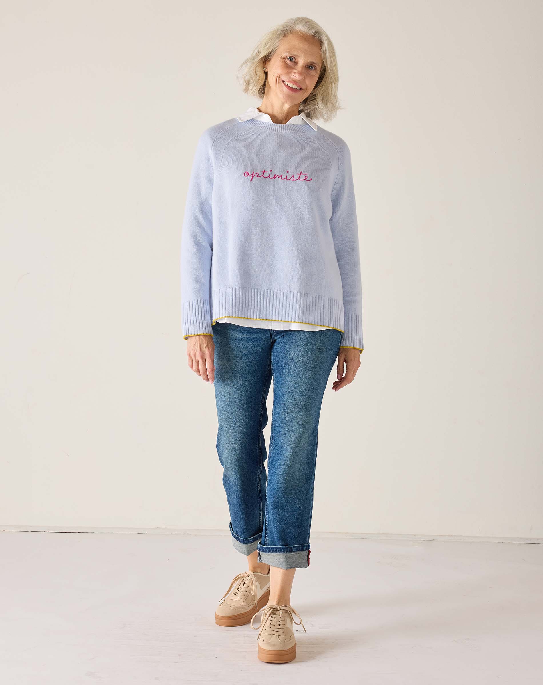 Woman wearing a light blue sweater with 'Optimist' text and blue jeans on a white background
