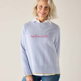 Woman wearing a light blue sweater with 'optimiste' text, standing against a plain background