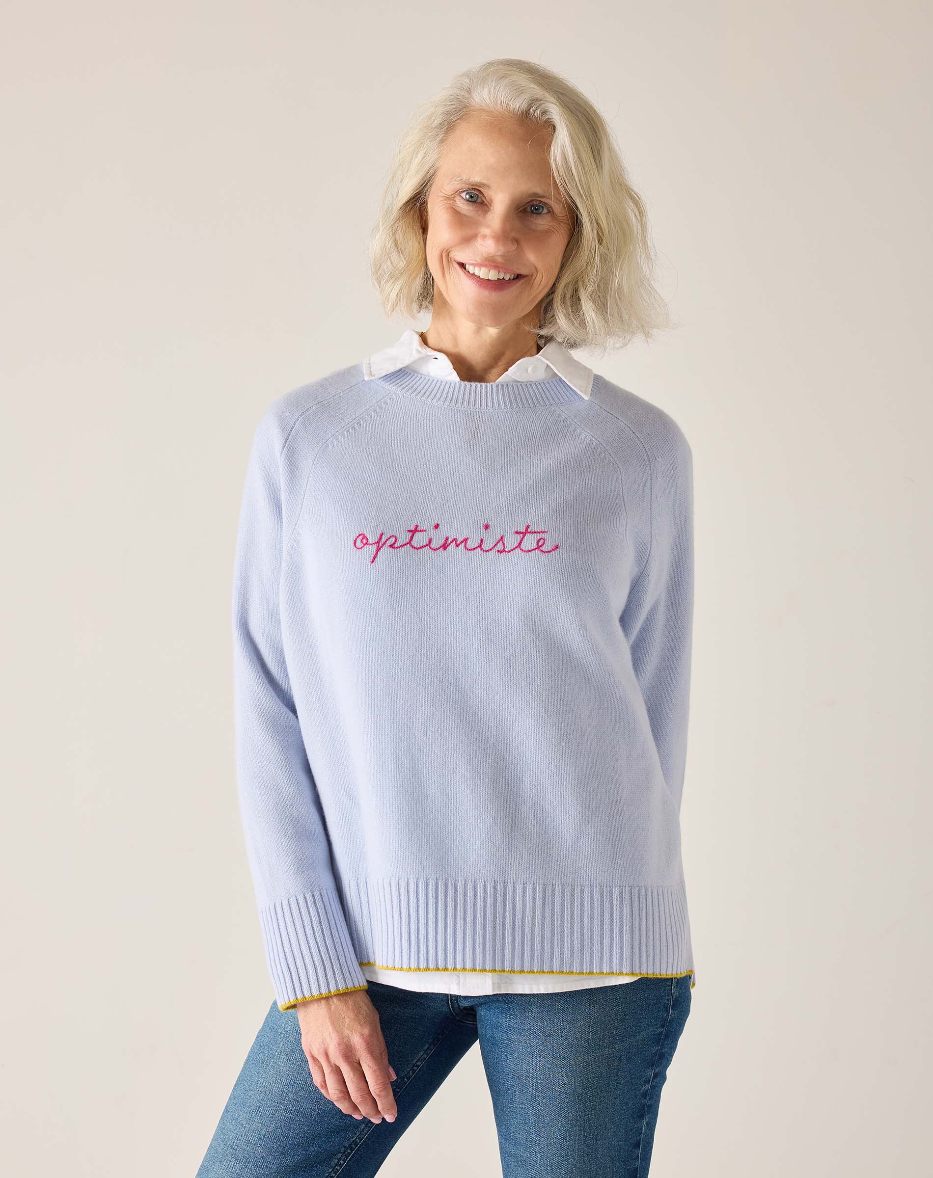 Sky blue crewneck knit sweater with 'optimiste' embroidered in red and a white collar peeking through.