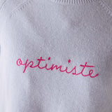 Light blue sweater with 'optimiste' embroidered in pink on a plain background