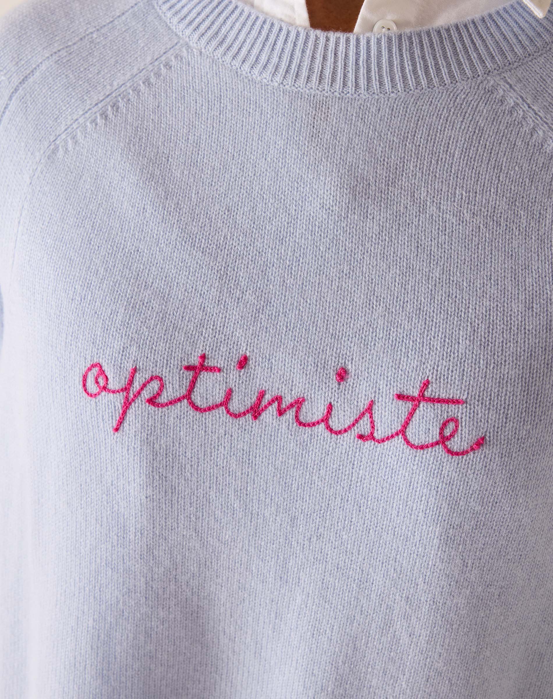 Light blue sweater with 'optimiste' embroidered in pink on a plain background