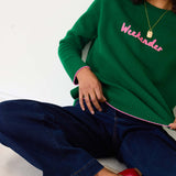 Green sweater with pink text 'Weekender' worn by a person on a white background