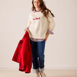 Women's travel-friendly off-white knit sweater featuring 'La Rebel' embroidery and a layering look with a red jacket and denim pants. Text: La Rebel