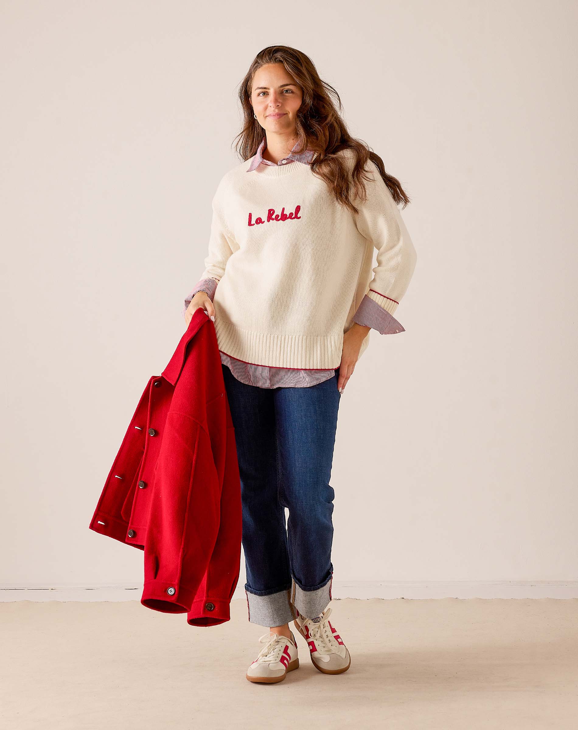 Women's travel-friendly off-white knit sweater featuring 'La Rebel' embroidery and a layering look with a red jacket and denim pants. Text: La Rebel