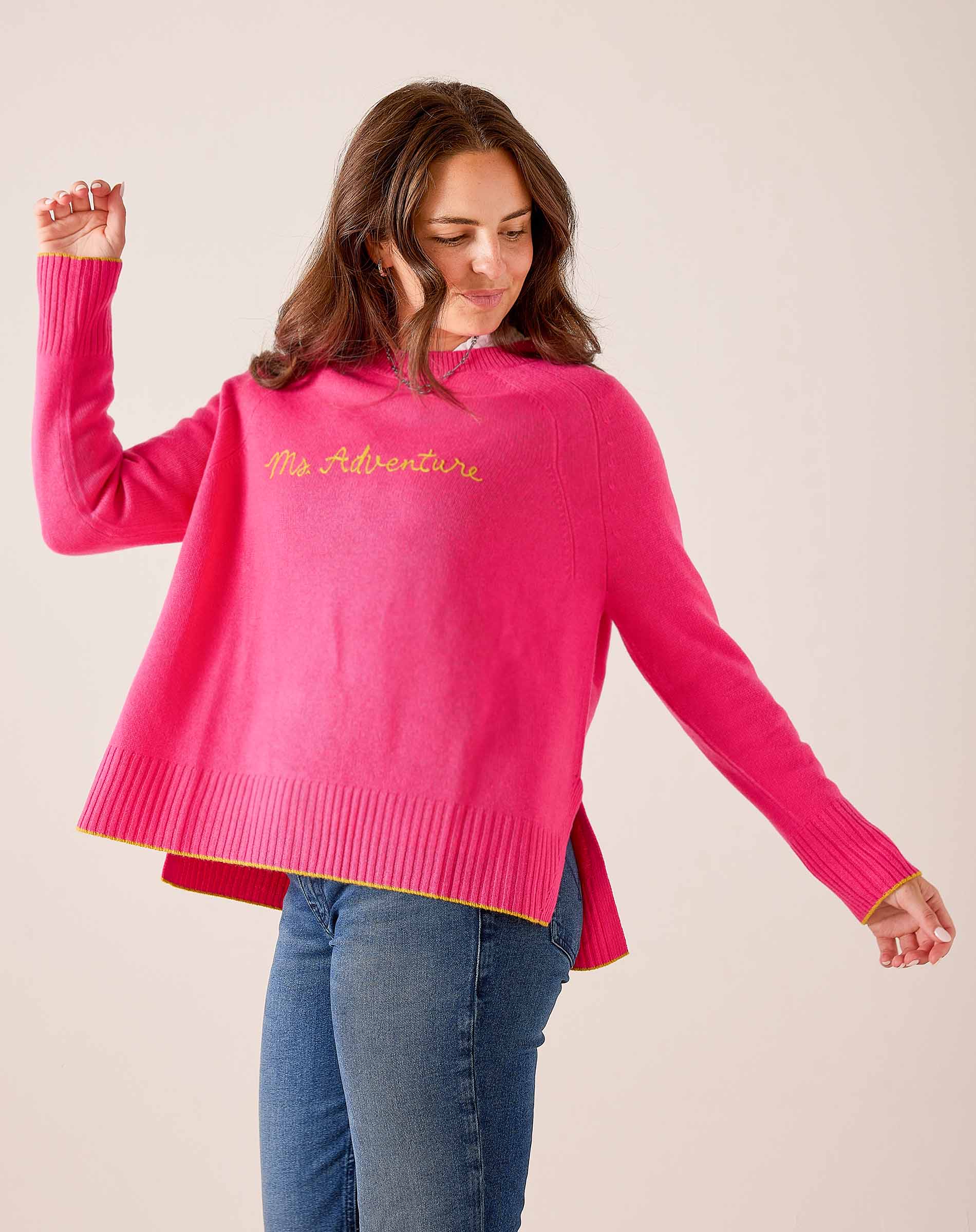 Hot pink oversized knit sweater with ribbed cuffs and hem, featuring 'Ms. Adventure' in gold script. Text: Ms. Adventure