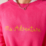 Hot pink knit sweater with a crew neck and 'Ms. Adventure' embroidered in yellow script. Text: Ms. Adventure