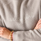 Gray sweater with 'salty girl' text worn by a person, with gold bracelet on wrist.