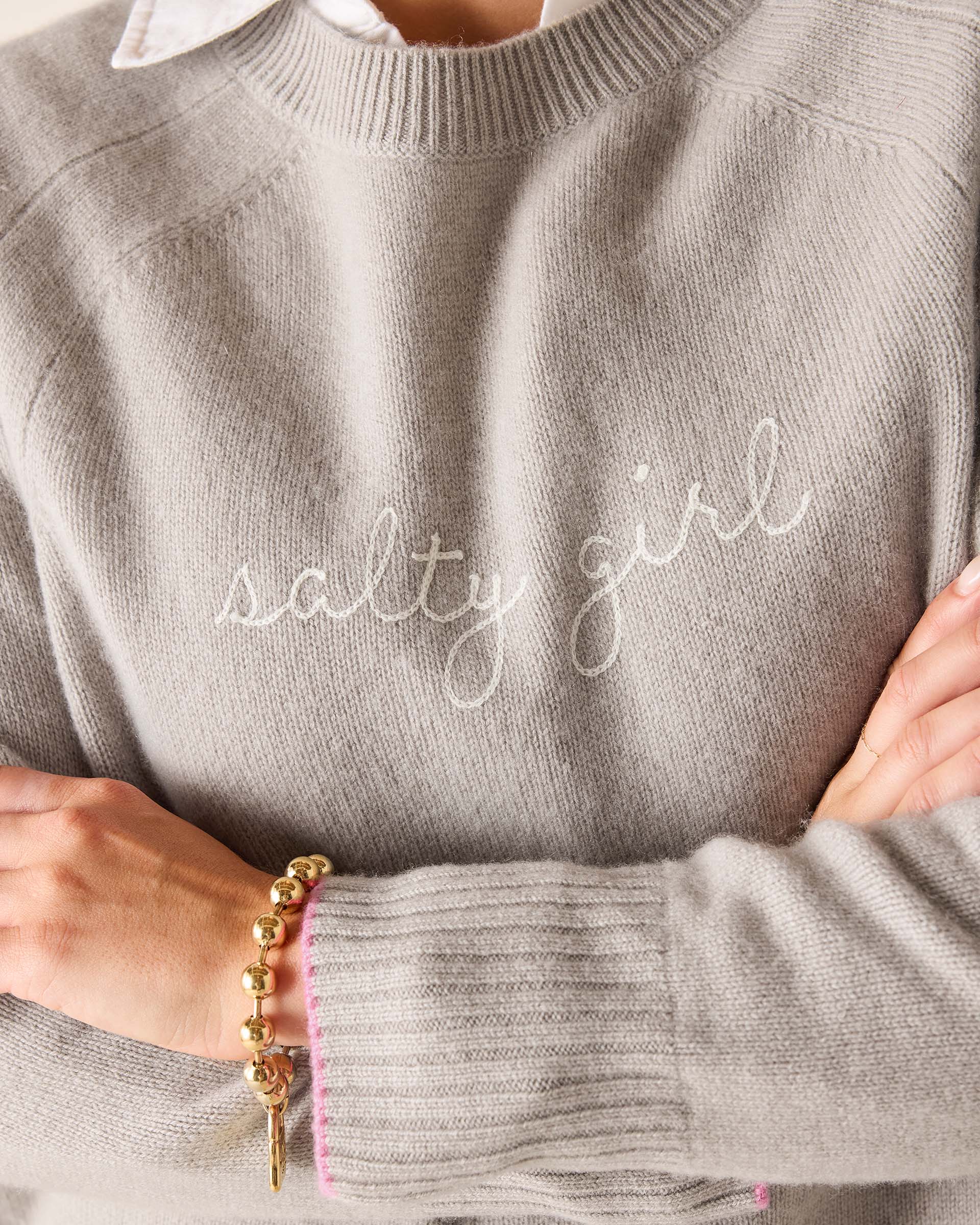 Gray sweater with 'salty girl' text worn by a person, with gold bracelet on wrist.