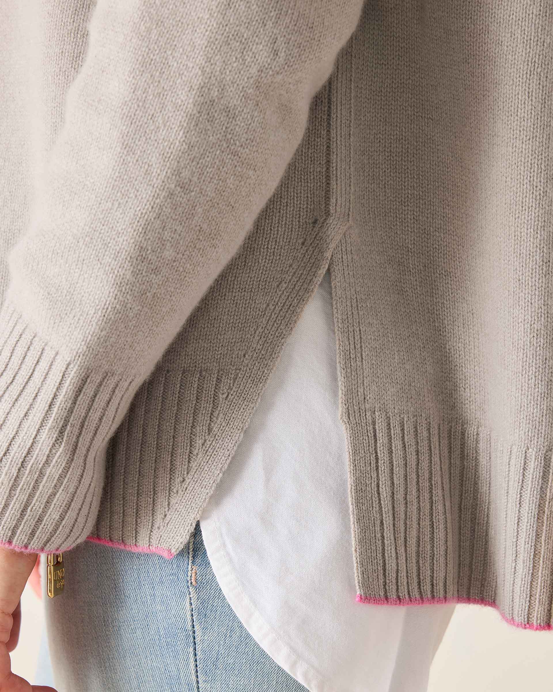 Close-up of a person wearing a beige sweater with pink trim, white shirt, and blue jeans.