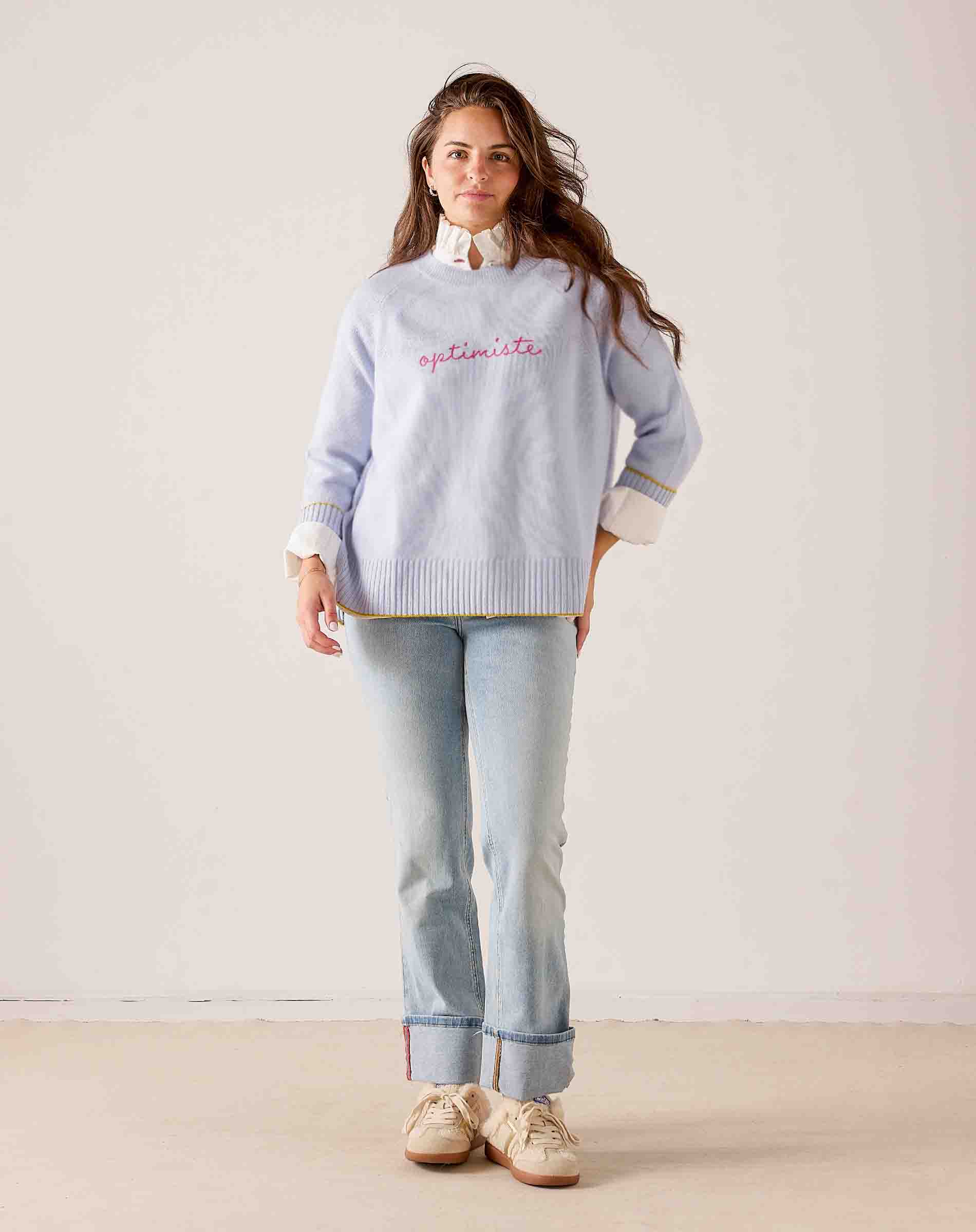Women's travel-friendly sky blue knit sweater with 'optimistic' embroidery and striped trim. Text: optimistic