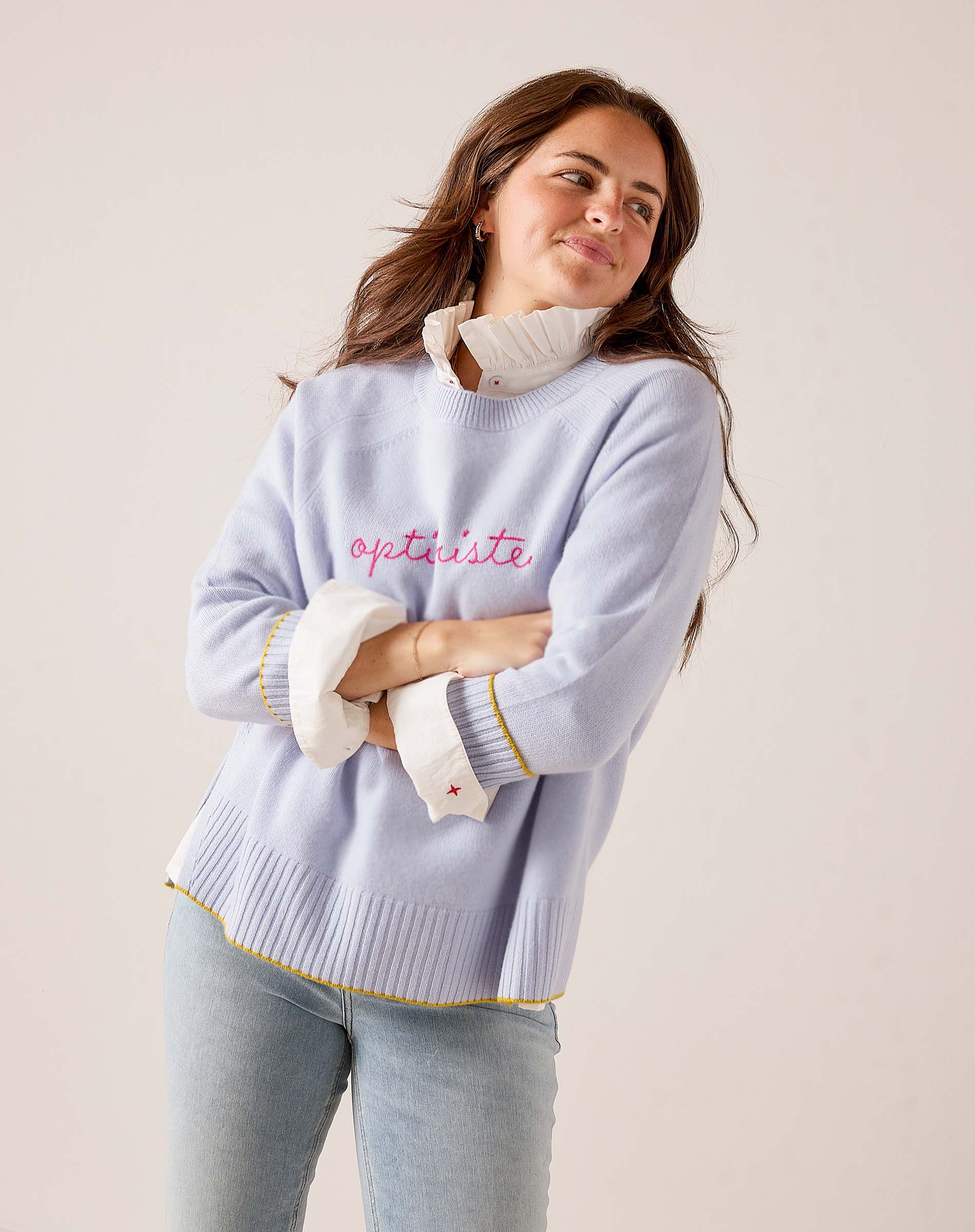 Sky blue oversized knit sweater with 'optimiste' script and yellow-trimmed ribbed details. Text: optimiste.