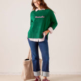 Women's travel-friendly emerald green knit sweater with 'Weekender' embroidered in pink. The model pairs the sweater with a white collared shirt, blue denim jeans, and red sneakers. Text: Weekender