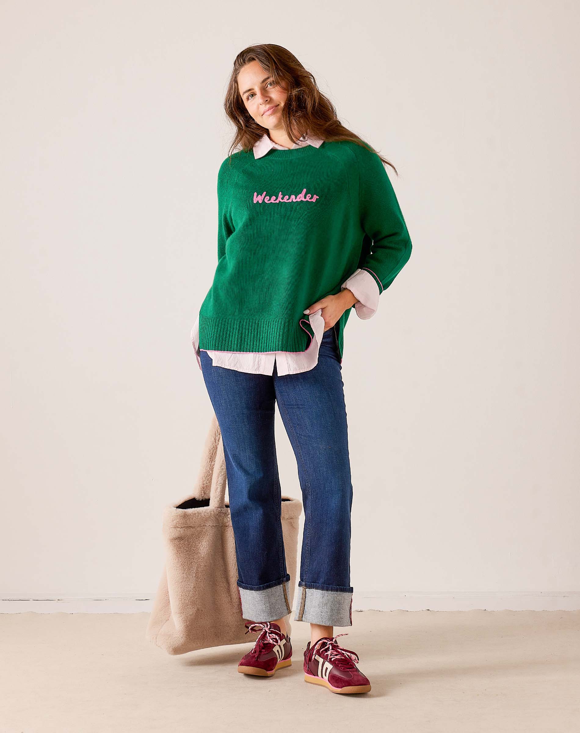 Women's travel-friendly emerald green knit sweater with 'Weekender' embroidered in pink. The model pairs the sweater with a white collared shirt, blue denim jeans, and red sneakers. Text: Weekender