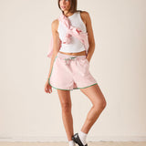 Woman wearing a white sleeveless top and pink shorts with a pink scarf, standing against a plain background.