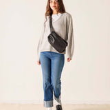 Woman wearing a gray sweater, blue jeans, and carrying a black crossbody bag against a white background