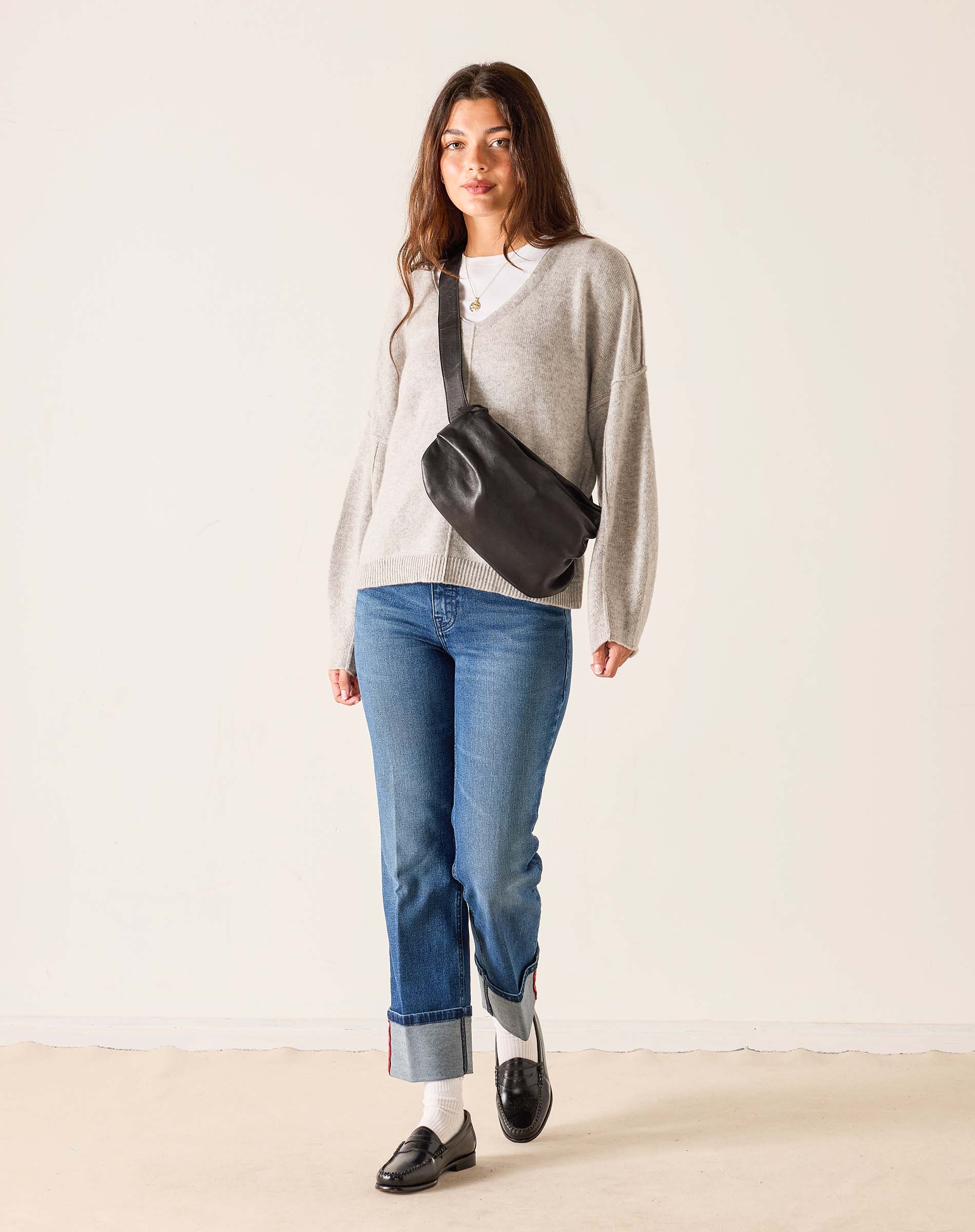 Woman wearing a gray sweater, blue jeans, and carrying a black crossbody bag against a white background