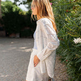 Woman wearing a light-colored outfit standing outdoors with greenery in the background
