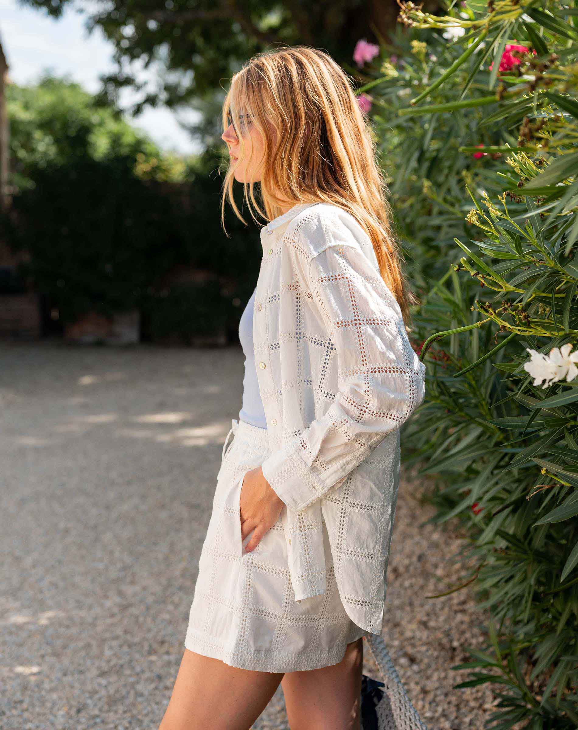 Woman wearing a light-colored outfit standing outdoors with greenery in the background