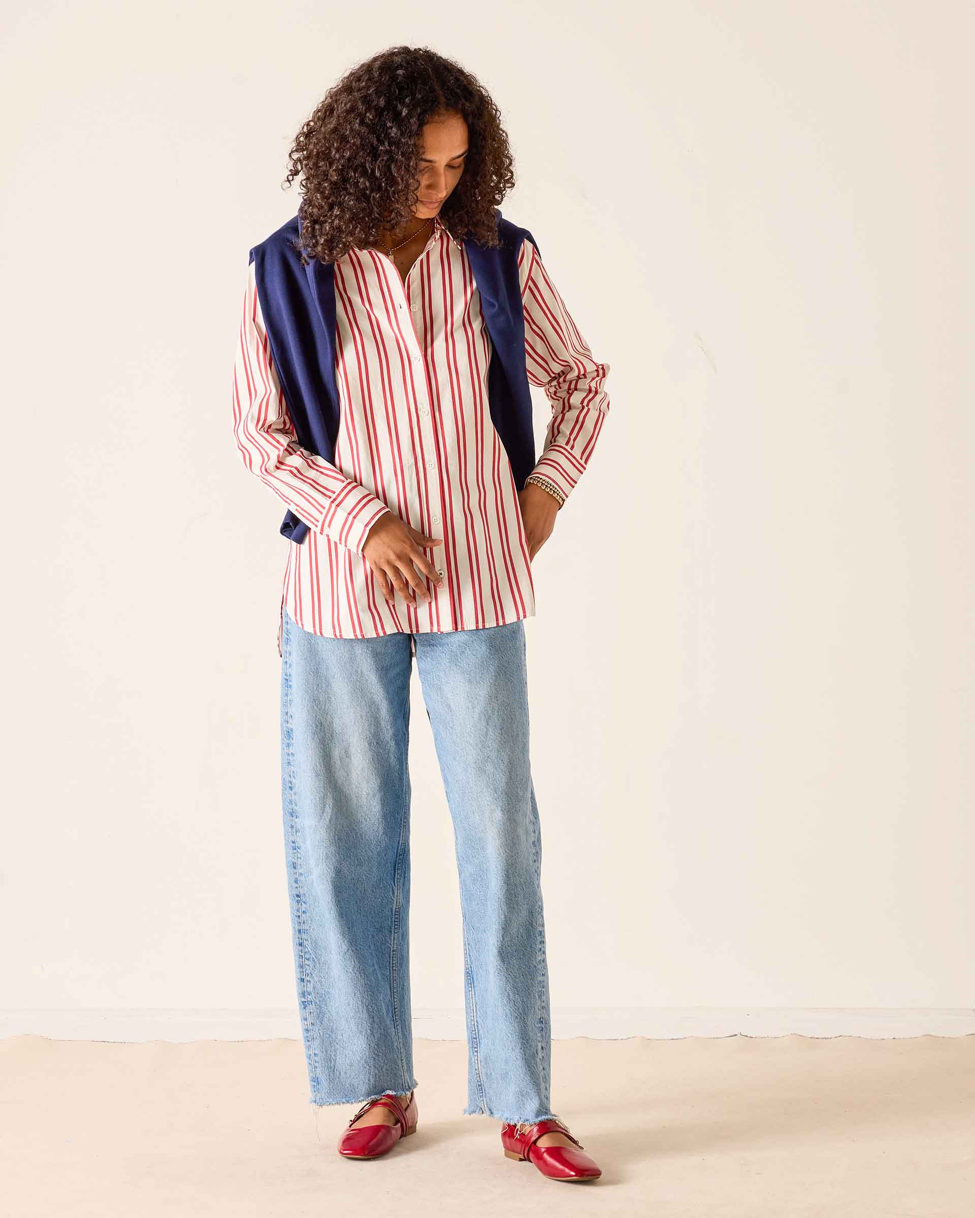 Woman wearing a striped shirt, blue jeans, and red shoes against a plain background
