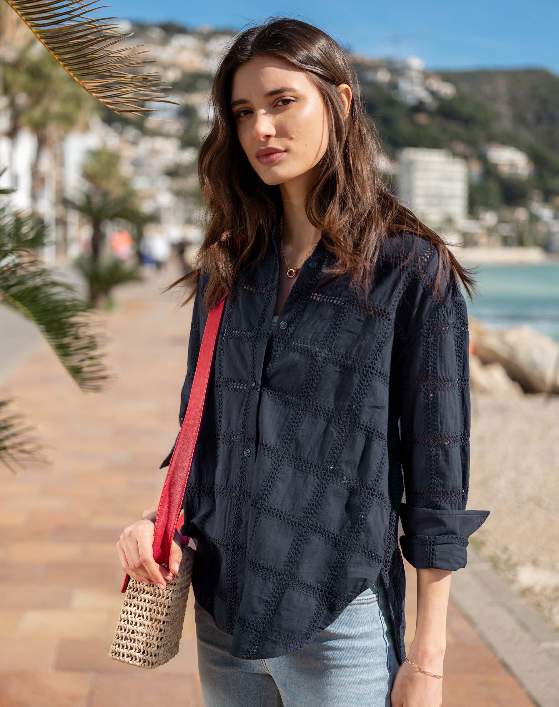 Woman standing on a coastal path holding a red bag, wearing a dark blue shirt and light blue jeans.
