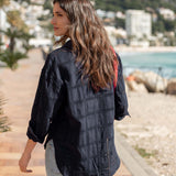 Woman wearing a black jacket with a plaid pattern, standing on a path by a waterfront.