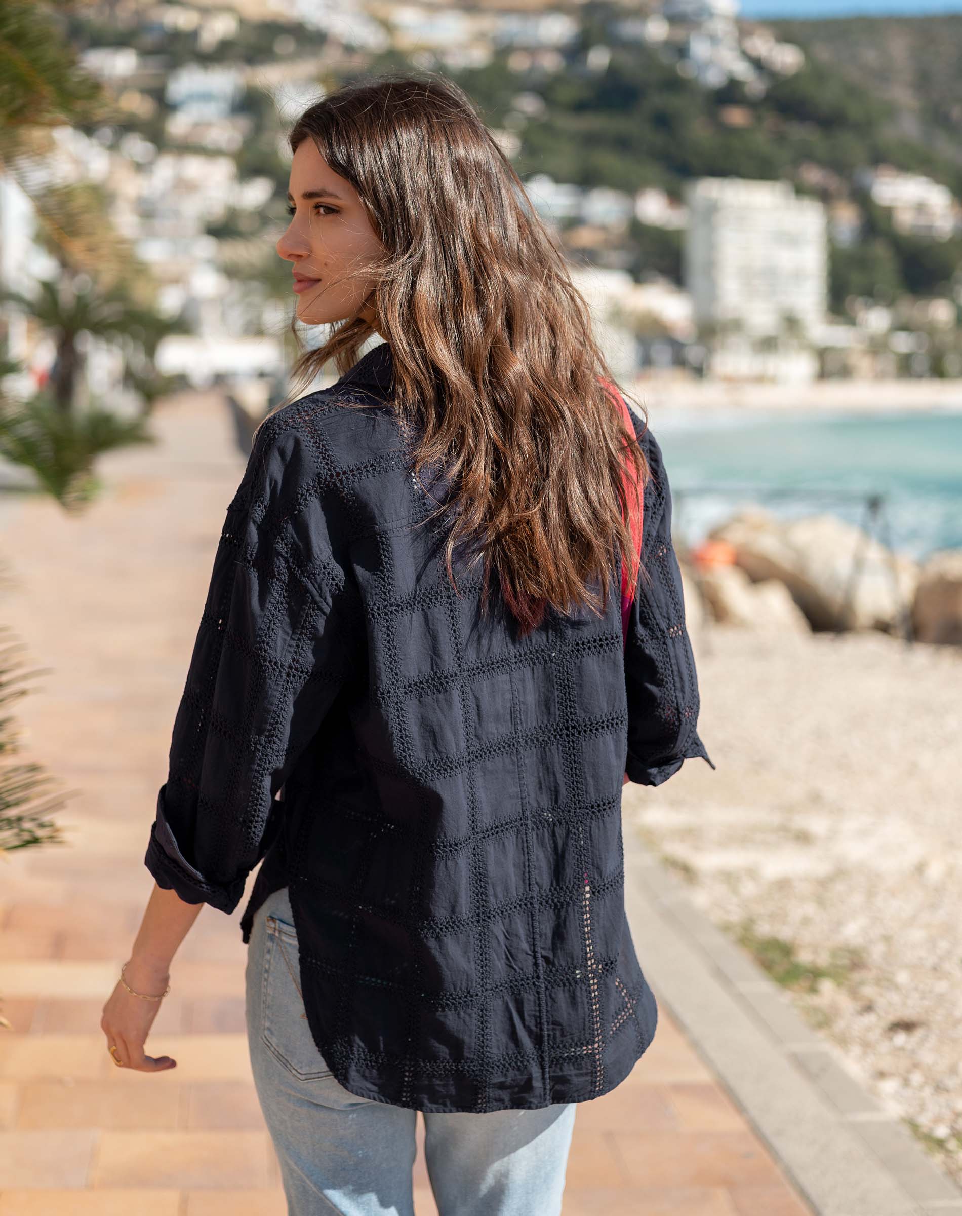 Woman wearing a black jacket with a plaid pattern, standing on a path by a waterfront.