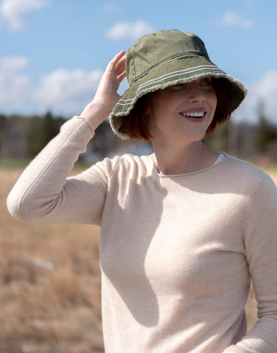female wearing green bucket hat and neutral sweater standing outside in a field on a sunny day