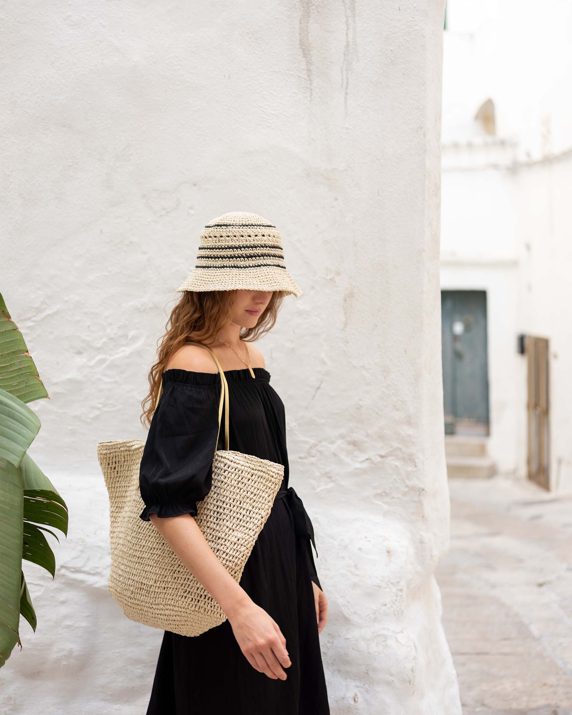 woman in italy against a white wall wearing calypso bucket hat in a black dress with tote bag on