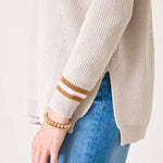 Beige sweater with gold and white striped cuffs worn by a person on a plain background