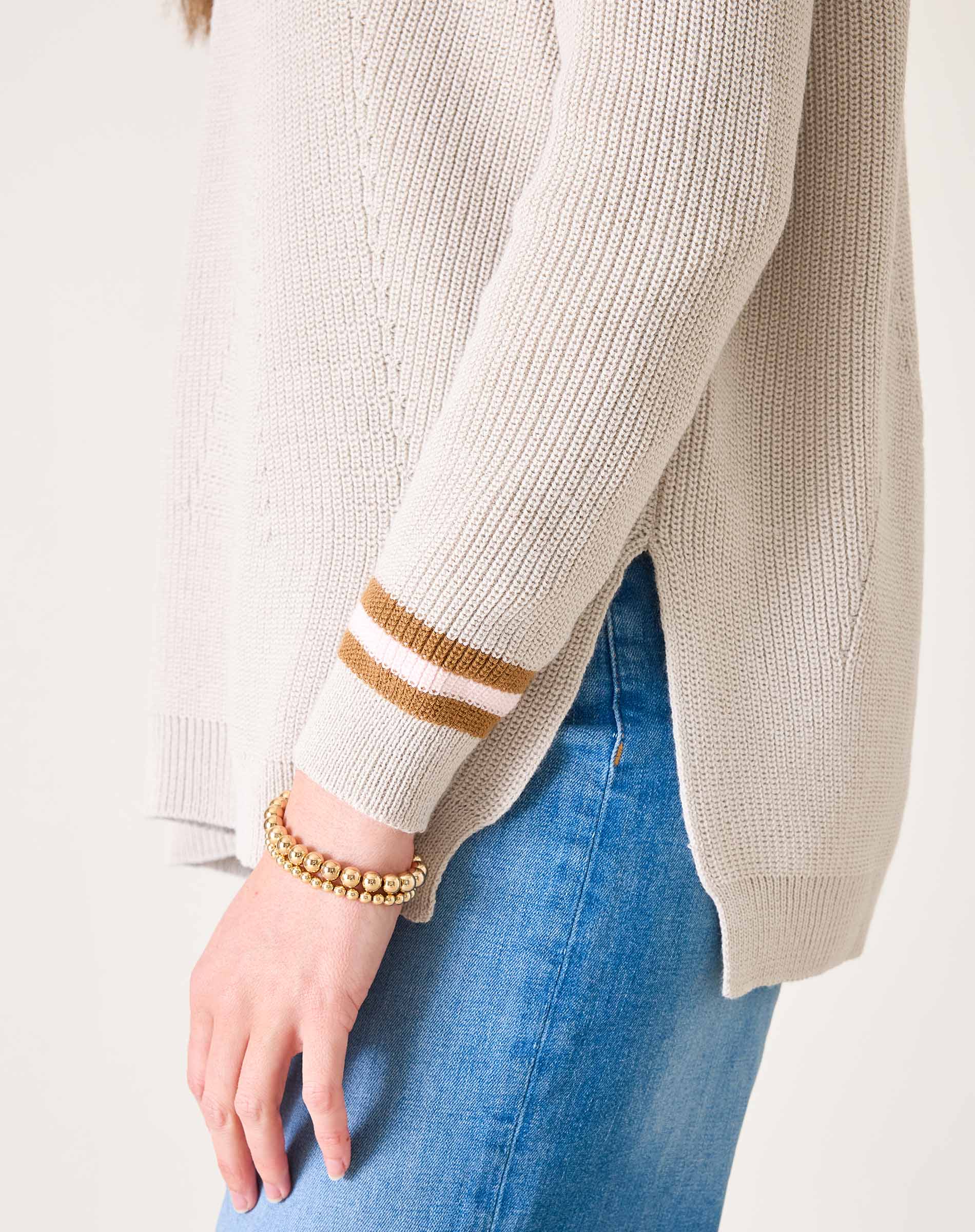 Beige sweater with gold and white striped cuffs worn by a person on a plain background