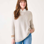 Woman wearing a beige sweater with gold trim and blue jeans on a white background
