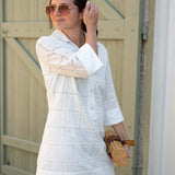 Woman wearing a white dress with a textured pattern, holding sunglasses and a woven bag, standing in front of a wooden door.