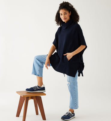 woman wearing mersea cape poncho sweater in navy blue with leg up on stool