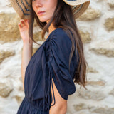 Woman wearing a navy dress and straw hat against a stone wall.