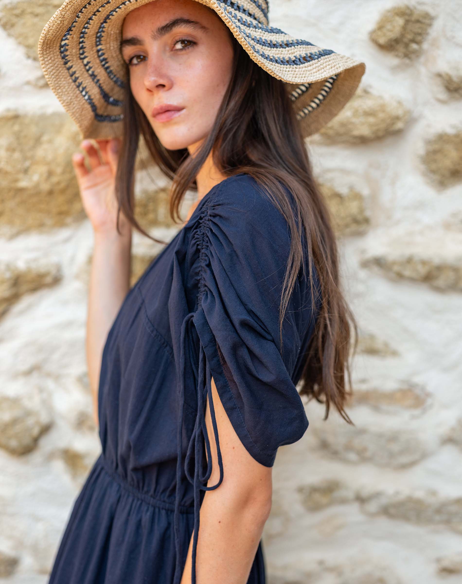 Woman wearing a navy dress and straw hat against a stone wall.