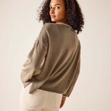 Woman wearing a brown sweater and white pants against a plain background