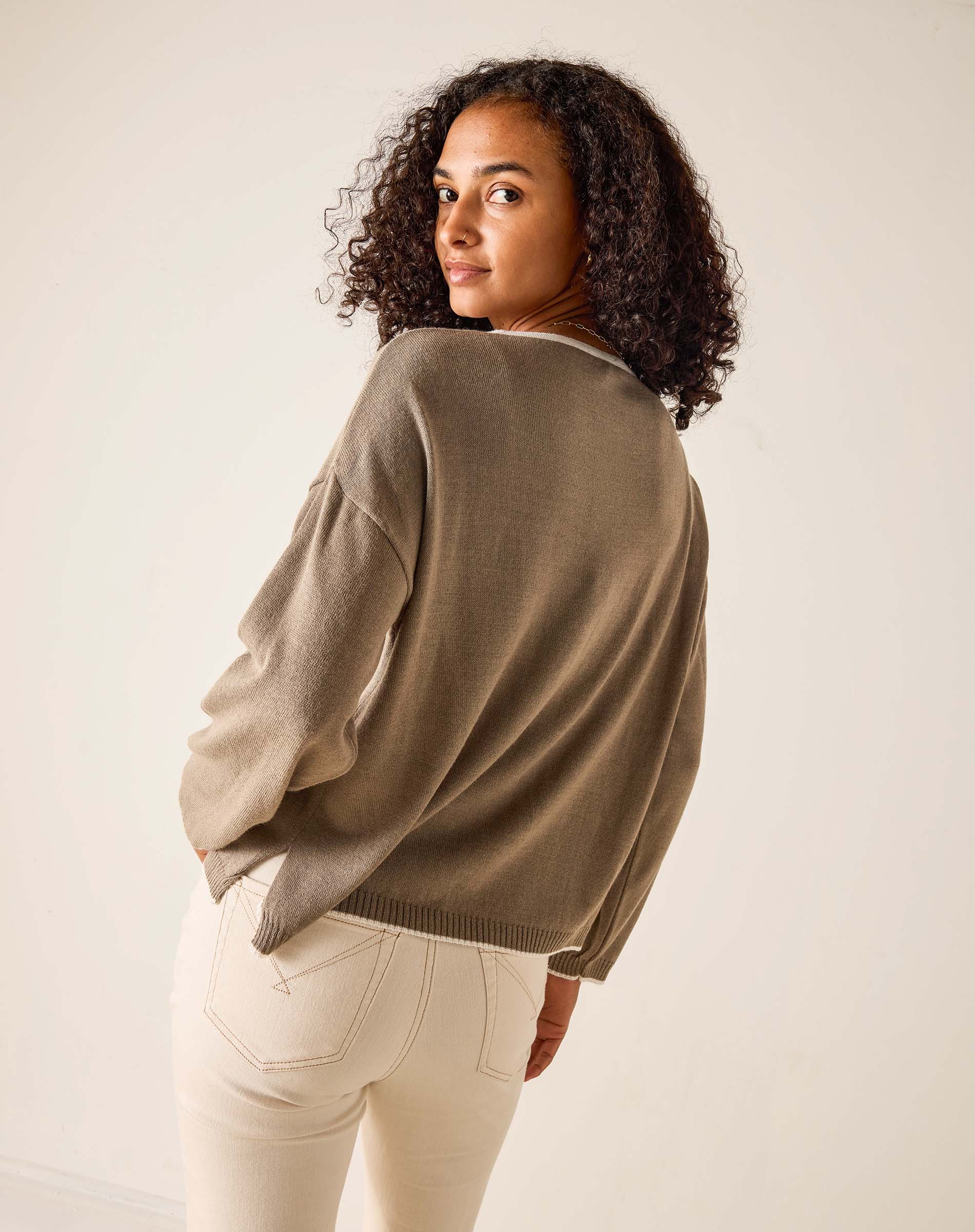 Woman wearing a brown sweater and white pants against a plain background
