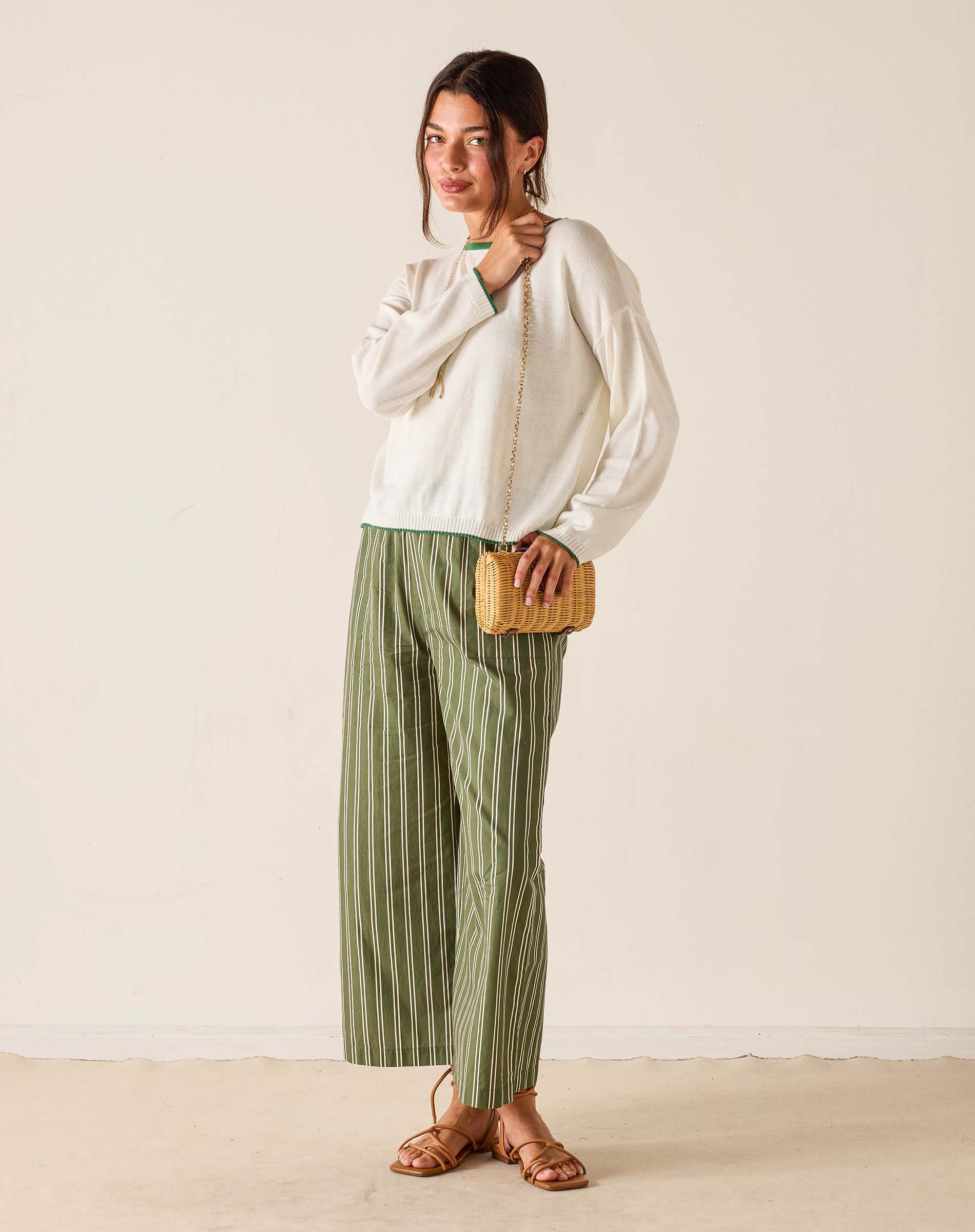 Woman wearing a white blouse and green striped pants, holding a wooden clutch against a plain background