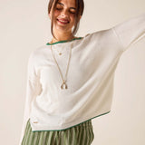 Woman wearing a white sweater with green trim and striped pants, standing against a plain background.
