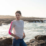 woman wearing mersea carmel cashmere sweater in champagne color with hot pink stripes down sleaves standing on rocks in front of body of water