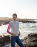 woman wearing mersea carmel cashmere sweater in champagne color with hot pink stripes down sleaves standing on rocks in front of body of water