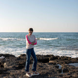 woman wearing mersea carmel cashmere sweater in champagne color with hot pink stripes down sleaves standing on rocks in front of body of water