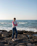 woman wearing mersea carmel cashmere sweater in champagne color with hot pink stripes down sleaves standing on rocks in front of body of water