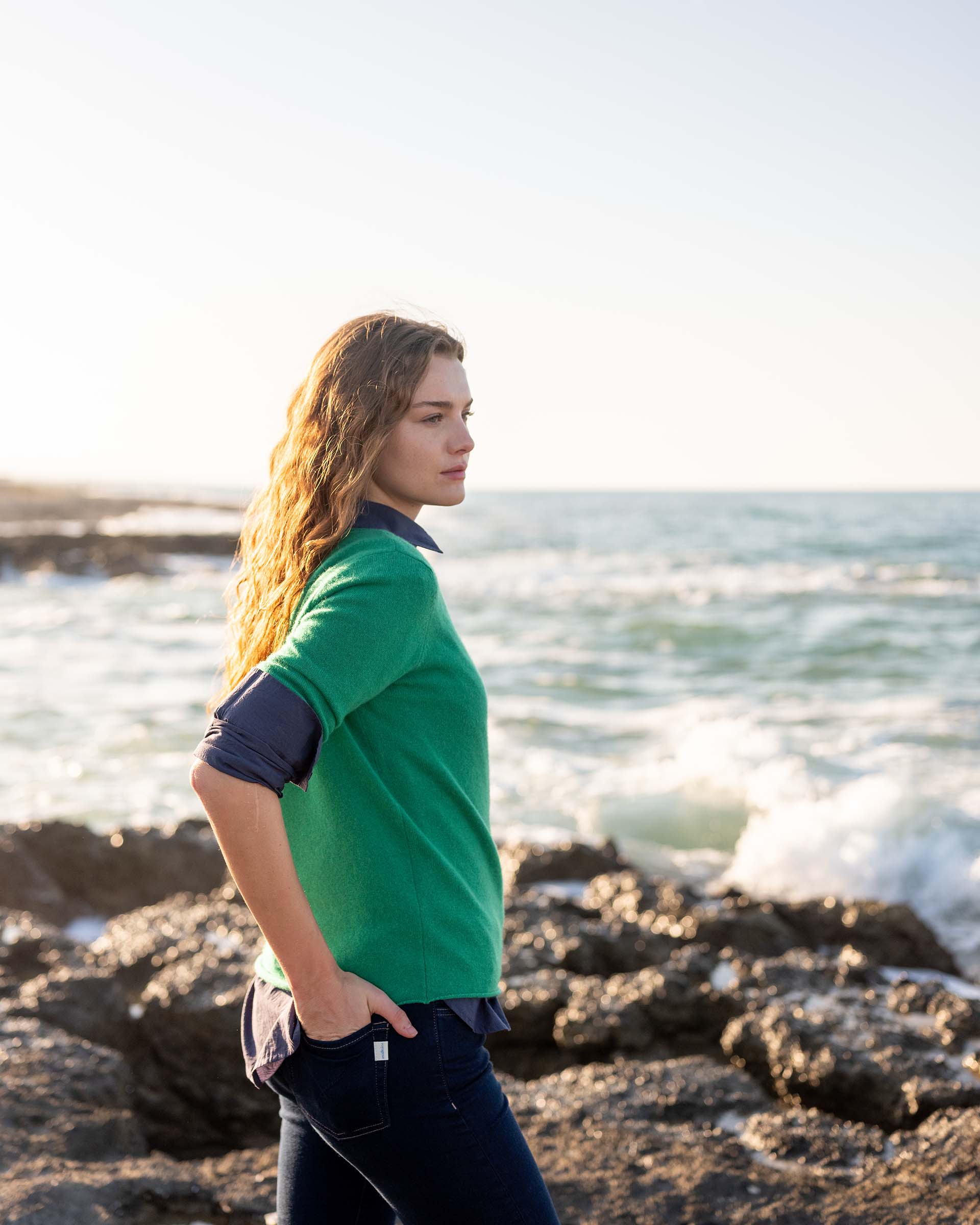 profile of woman wearing mersea carmel sweater in jade standing near water on rocks