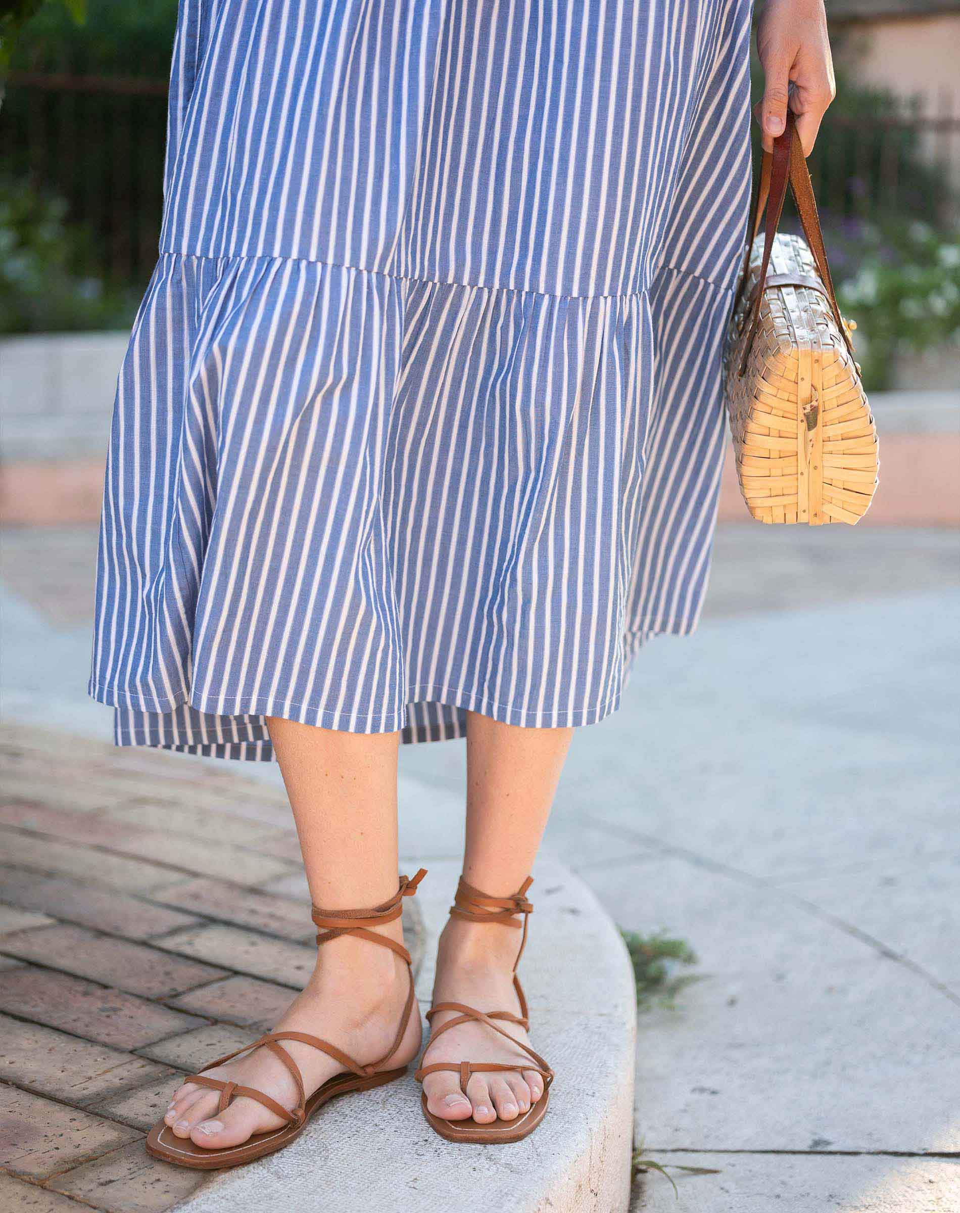 Person wearing a blue and white striped dress with brown sandals and a straw bag.